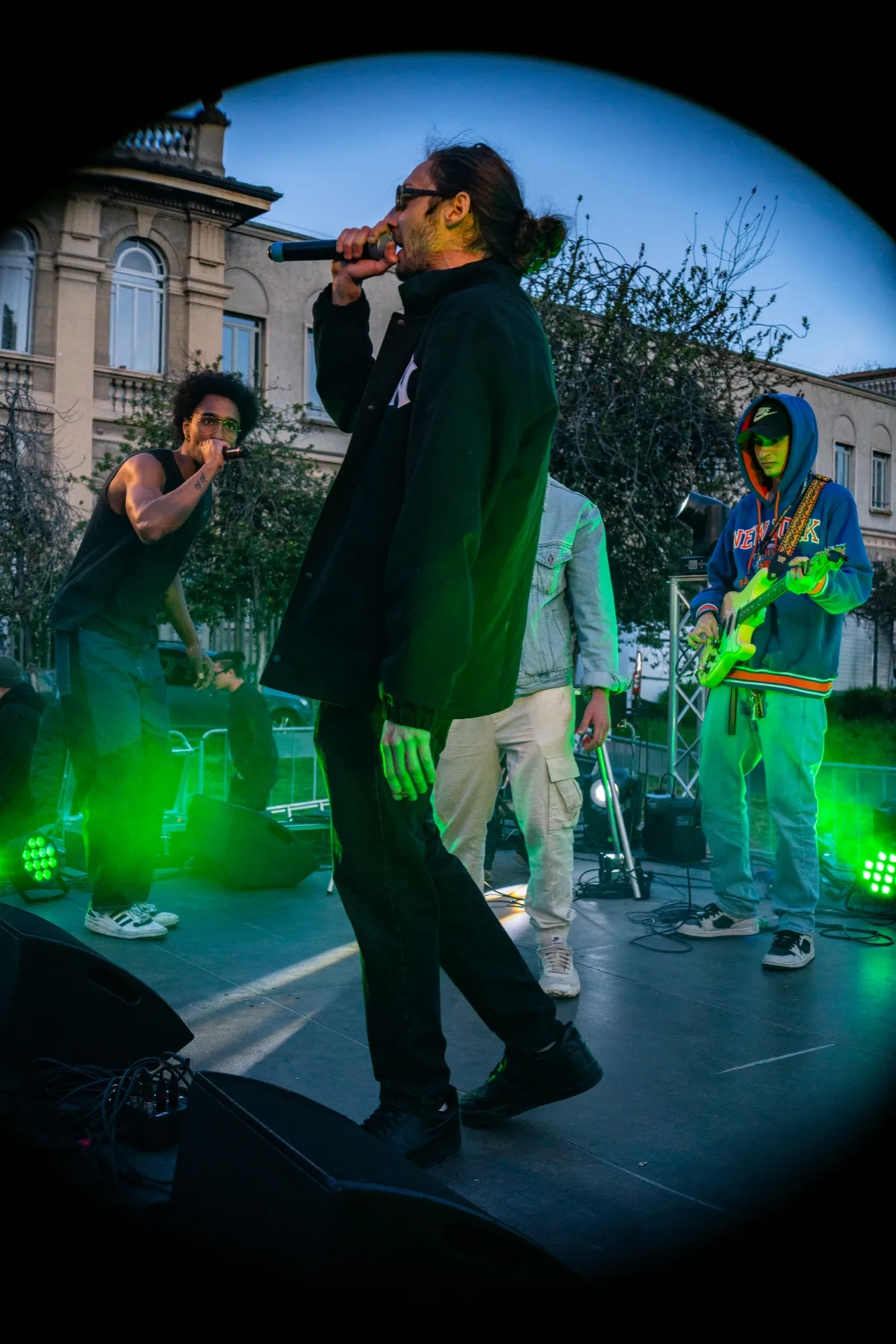 Il cantante con il microfono si muove sul palco all'aperto, due musicisti alle spalle e un palazzo storico sullo sfondo al crepuscolo